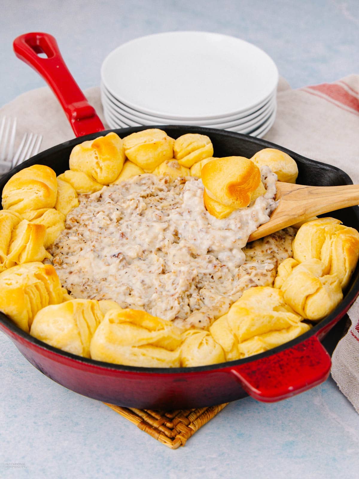 A red cast iron skillet filled with creamy sausage gravy in the center, surrounded by golden baked biscuits. A wooden spoon is dipping into the gravy, and a stack of white plates is in the background.