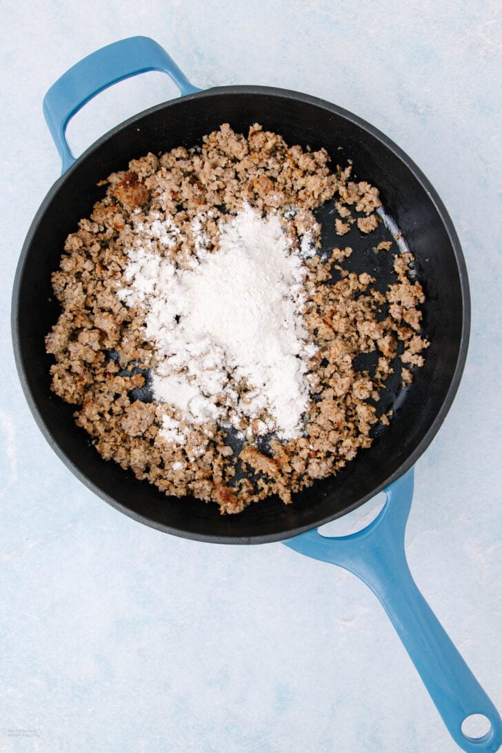A blue skillet filled with cooked ground sausage and a mound of white flour in the center, set on a light blue surface.