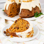 A slice of carrot cake with cream cheese frosting and walnuts sits on a white plate. In the background, the rest of the frosted bundt cake is displayed on a separate plate.