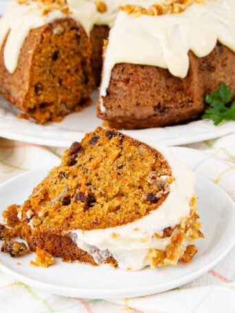 A slice of carrot cake with cream cheese frosting and walnuts sits on a white plate. In the background, the rest of the frosted bundt cake is displayed on a separate plate.
