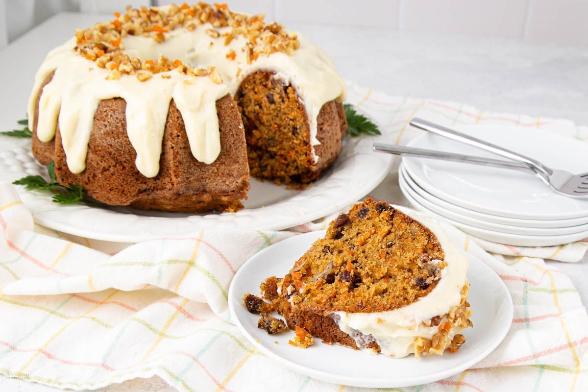 A bundt-shaped carrot cake with cream cheese frosting and chopped nuts on top sits on a plate. A slice is served on a smaller plate nearby, with forks and stacked plates in the background.
