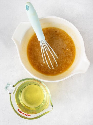 A mixing bowl with a light blue whisk in a brown liquid mixture, next to a glass measuring cup filled with oil, both on a white countertop.
