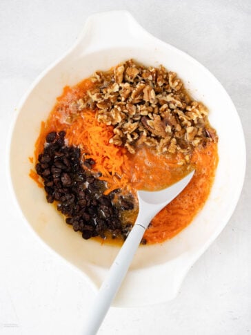 A white mixing bowl contains shredded carrots, chopped walnuts, and raisins, partially mixed with a wet batter. A gray and white mixing spoon rests inside the bowl.