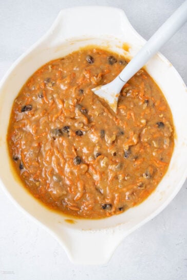 A white mixing bowl filled with thick carrot cake batter containing visible shredded carrots, raisins, and walnuts, with a white spatula resting inside the bowl.