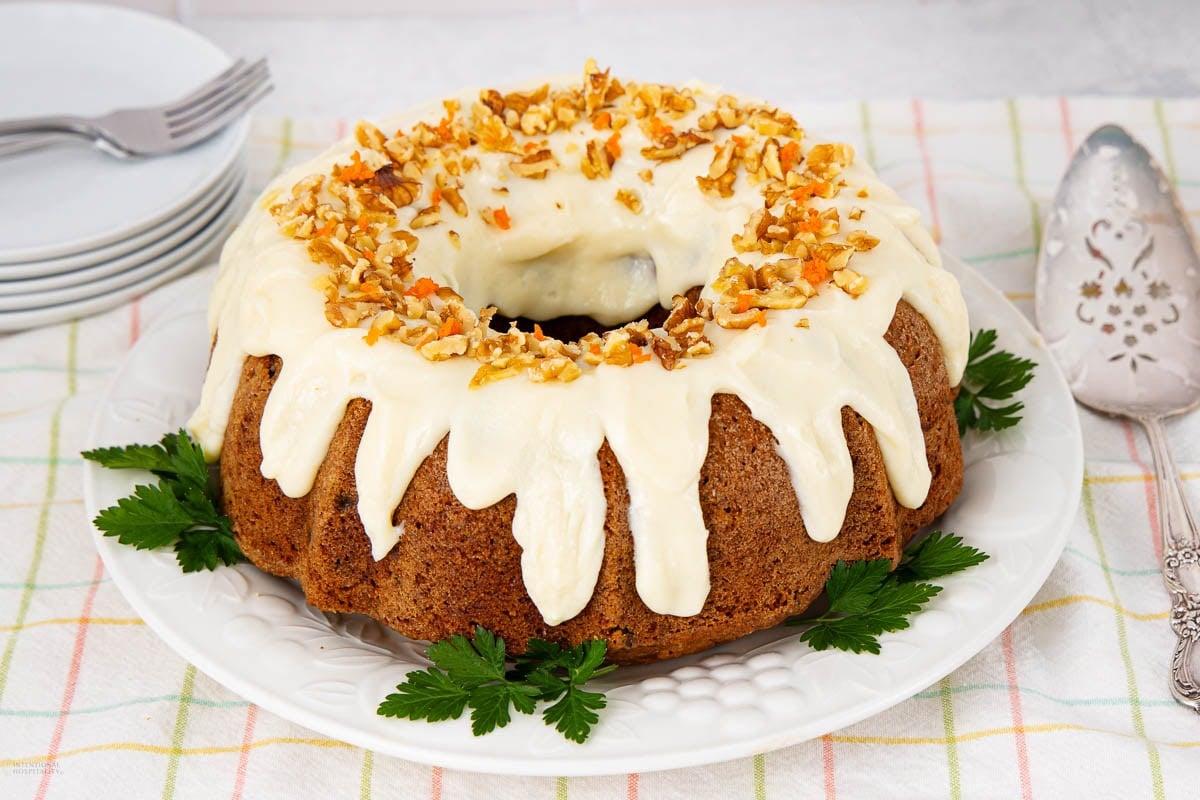 A round carrot cake with white icing drips and chopped nuts on top, served on a white plate with parsley garnish. Plates, forks, and a cake server are nearby on a checkered tablecloth.