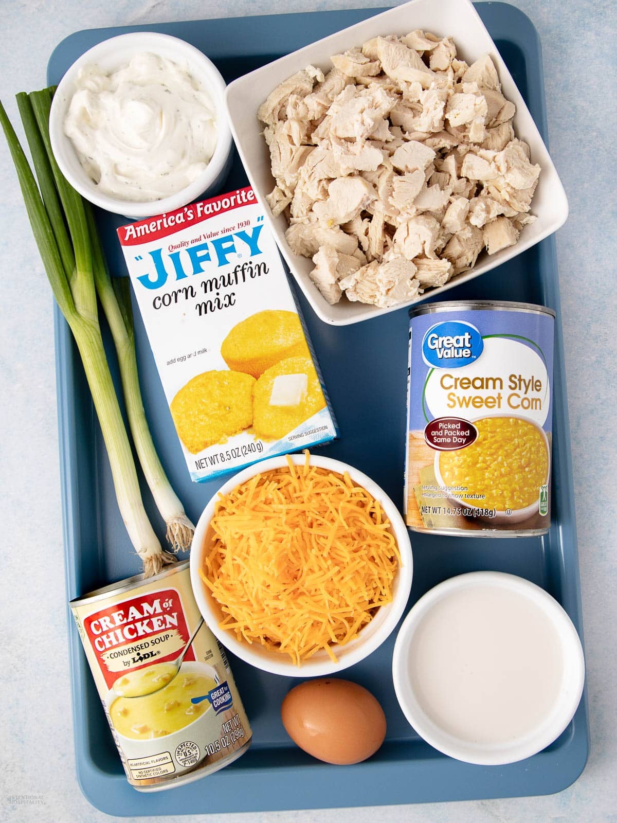 A blue tray holds green onions, sour cream, Jiffy corn muffin mix, chopped cooked chicken, shredded cheese, a can of cream-style corn, a can of cream of chicken soup, milk in a bowl, and an egg.