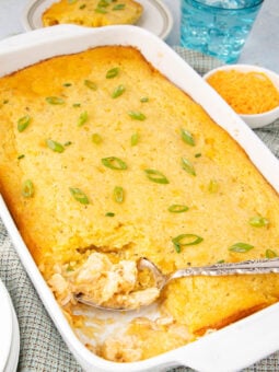 A white baking dish filled with golden cornbread casserole, topped with chopped green onions. A serving spoon is scooping out a portion, showing a creamy filling. Nearby are plates, a bowl of shredded cheese, and a glass of water.