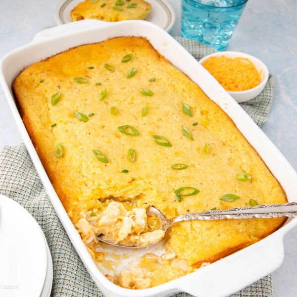 A white baking dish filled with golden cornbread casserole, topped with chopped green onions. A serving spoon is scooping out a portion, showing a creamy filling. Nearby are plates, a bowl of shredded cheese, and a glass of water.