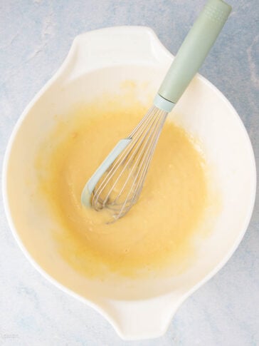 A white mixing bowl containing pale yellow batter with a whisk resting inside, placed on a light blue surface.