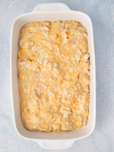 A white baking dish filled with an unbaked mixture of shredded cheese, chopped chicken, and a creamy sauce on a light blue surface.