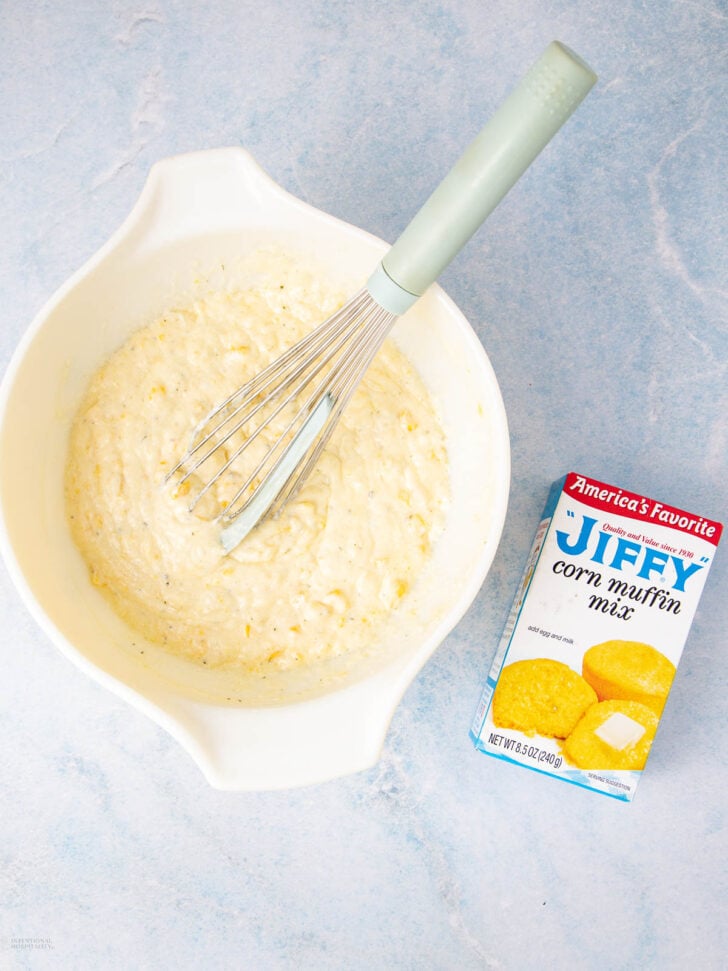A mixing bowl with corn muffin batter and a whisk beside a box of Jiffy corn muffin mix on a light blue surface.
