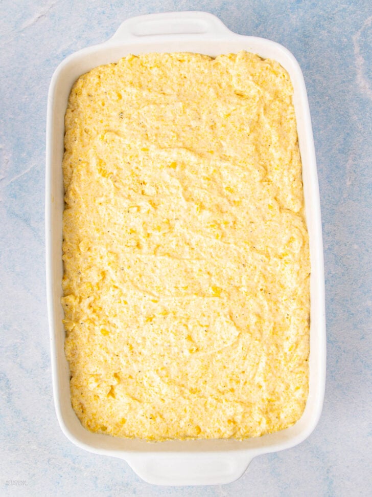 A white rectangular baking dish filled with an even layer of unbaked cornbread batter, set on a light blue surface.