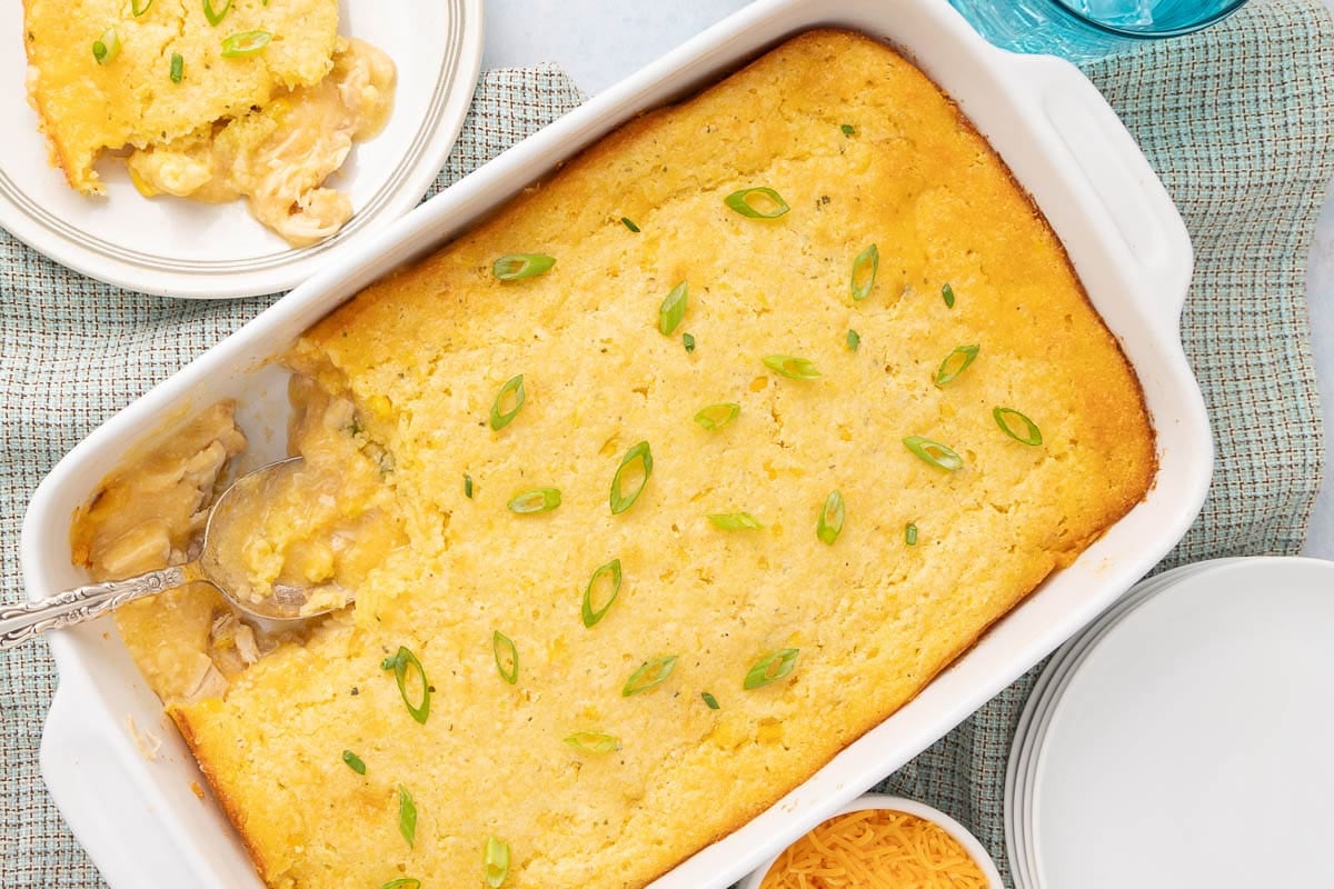 A baked casserole topped with golden cornbread and sliced green onions sits in a white dish. A serving has been scooped out onto a plate, revealing a creamy filling underneath. A spoon rests in the casserole.