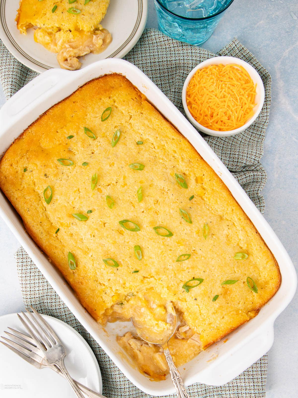 A baked casserole topped with golden cornbread and sliced green onions, served in a white dish. A portion is removed, showing a creamy filling. Nearby are plates, forks, a bowl of shredded cheese, and a glass of water.