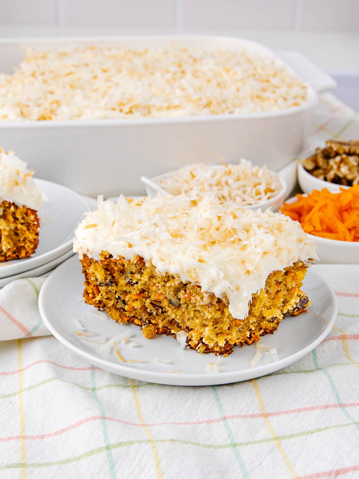 A slice of carrot cake topped with cream cheese frosting and shredded coconut sits on a plate, with the rest of the cake in a baking dish and bowls of walnuts and shredded carrots in the background.
