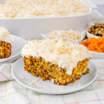 A slice of carrot cake with cream cheese frosting and shredded coconut on top sits on a white plate, with bowls of shredded coconut, walnuts, and grated carrots in the background.