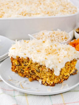 A slice of carrot cake with cream cheese frosting and shredded coconut on top sits on a white plate, with bowls of shredded coconut, walnuts, and grated carrots in the background.