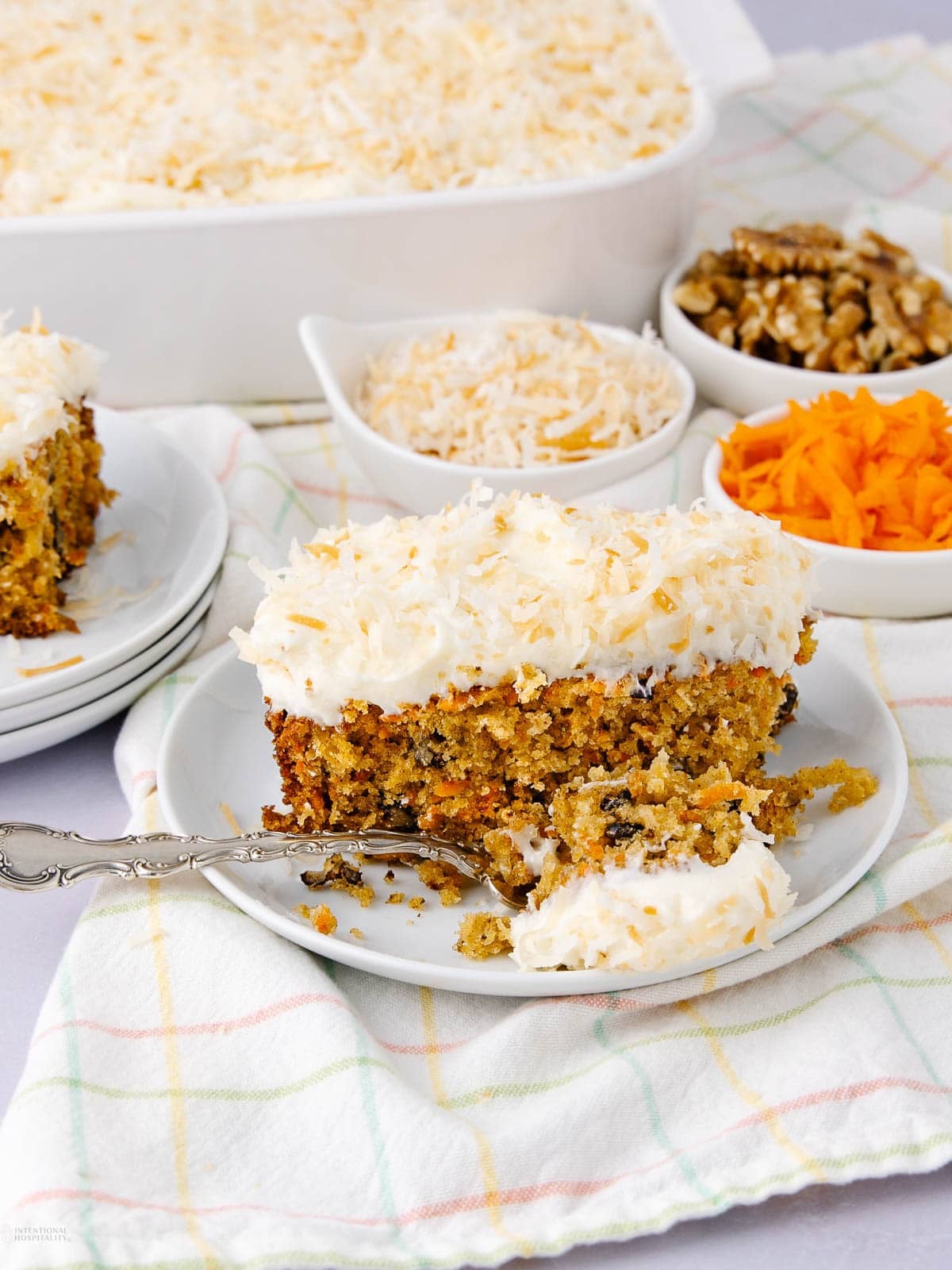 A slice of carrot cake with creamy white frosting and shredded coconut on top sits on a plate with a fork. Small bowls of shredded coconut, walnuts, and grated carrots are in the background.
