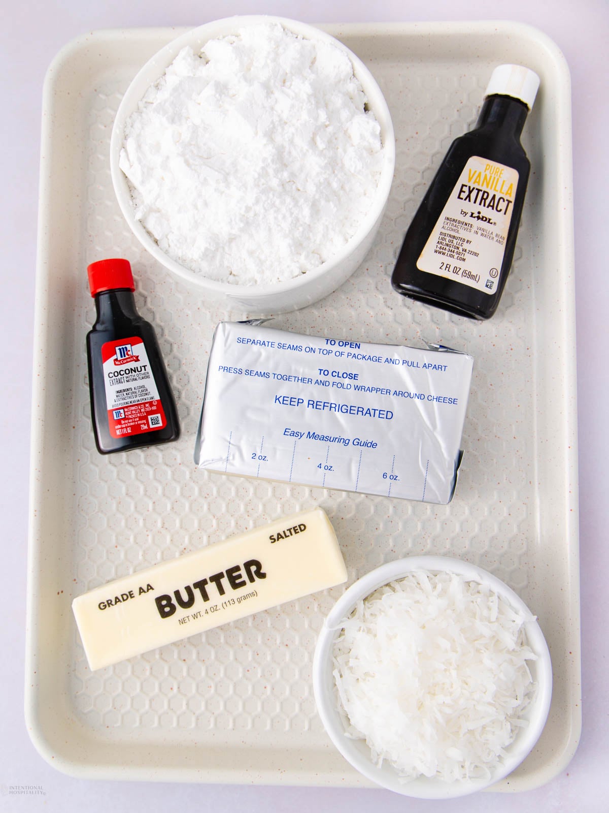A tray with powdered sugar in a bowl, a bottle of vanilla extract, a bottle of coconut extract, a stick of salted butter, a block of cream cheese, and a bowl of shredded coconut.
