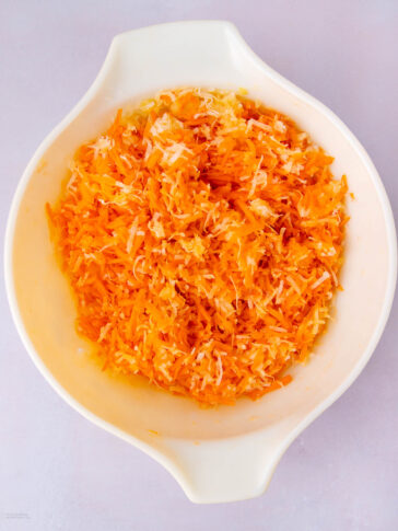 A white bowl filled with shredded orange carrots and white coconut, placed on a light surface.