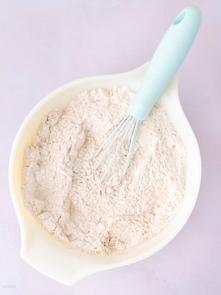 A white mixing bowl filled with flour and a blue-handled whisk resting on top, set on a light pink surface.