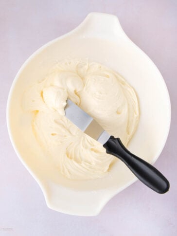A white mixing bowl filled with creamy, whipped frosting. A metal spatula with a black handle rests on top of the frosting inside the bowl.
