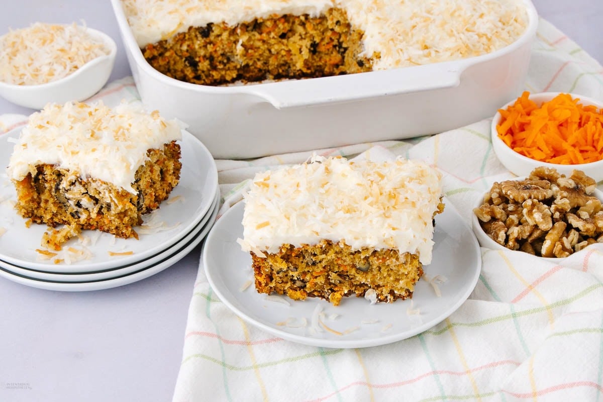 A slice of carrot cake with cream cheese frosting and toasted coconut sits on a plate, with more slices, grated carrots, walnuts, and a baking dish of cake in the background on a checkered cloth.