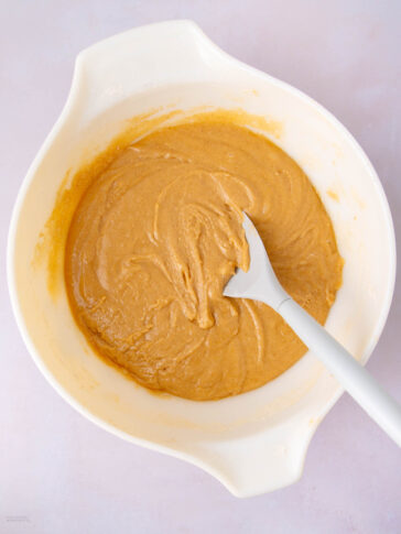 A white mixing bowl filled with light brown batter is shown on a pale surface. A white spatula rests in the bowl, partially covered with the smooth, swirled batter.