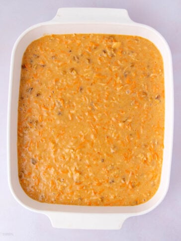 A white rectangular baking dish filled with an uncooked mixture of shredded carrots, ground meat, and sauce, ready to be baked.