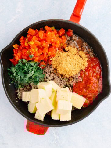 A skillet filled with cubed cheese, cooked ground beef, chopped tomatoes, cilantro, tomato sauce, and seasoning, all arranged in separate sections before mixing.