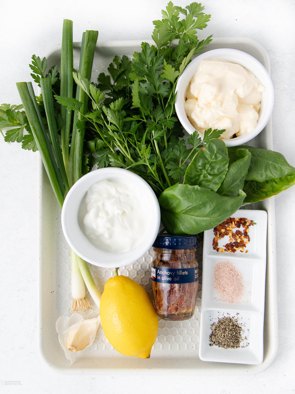 A tray holding fresh green onions, parsley, spinach, a lemon, garlic, a jar of anchovy fillets, bowls of mayonnaise and yogurt, and small dishes with red pepper flakes, salt, and black pepper.