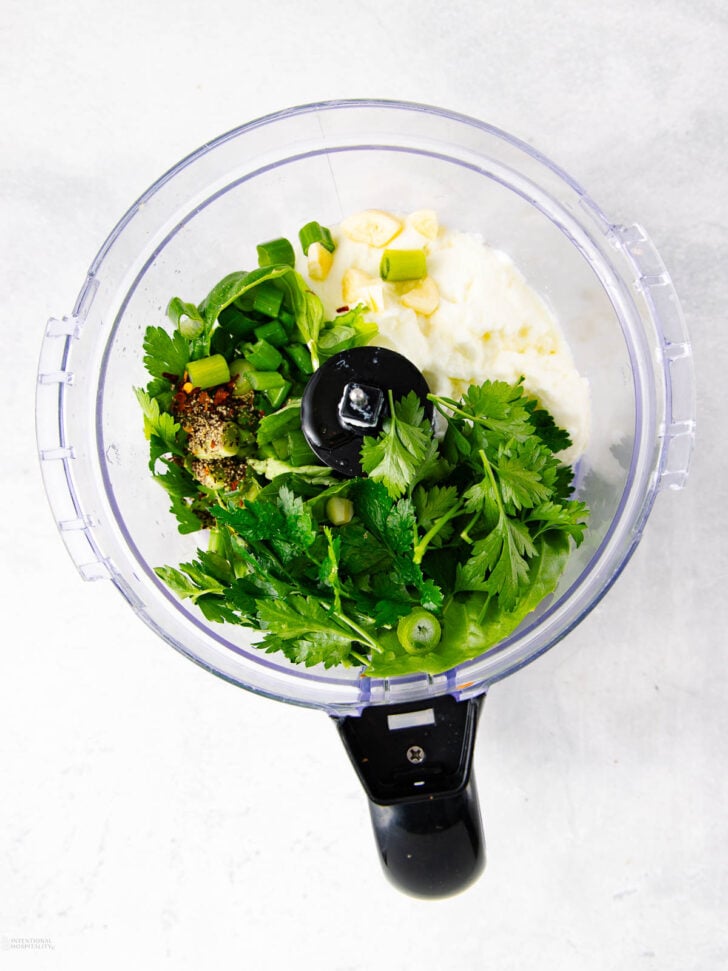 Overhead view of a food processor containing fresh parsley, green onions, garlic, spices, and a creamy white ingredient, ready to be blended on a light grey surface.