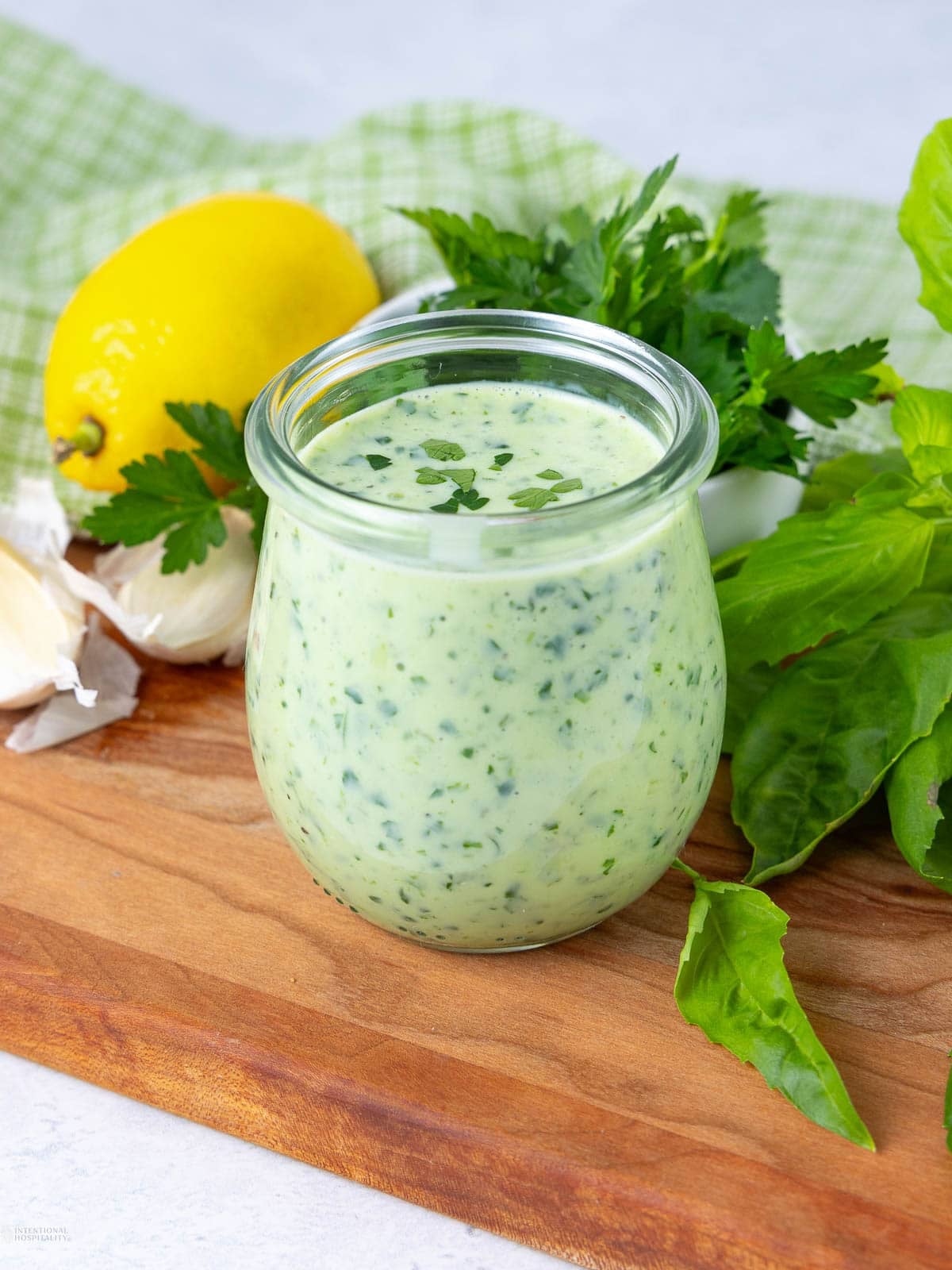 A glass jar filled with creamy green herb dressing sits on a wooden board, surrounded by fresh basil, parsley, garlic cloves, and a whole lemon, with a green checkered cloth in the background.