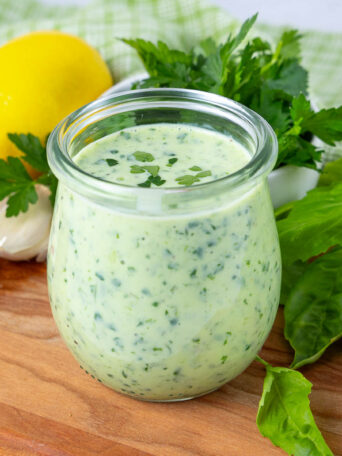 A glass jar filled with creamy green sauce sits on a wooden board, surrounded by fresh herbs, a lemon, and garlic cloves.