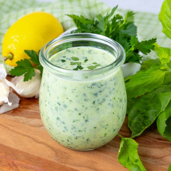 A glass jar filled with creamy green sauce sits on a wooden board, surrounded by fresh herbs, a lemon, and garlic cloves.