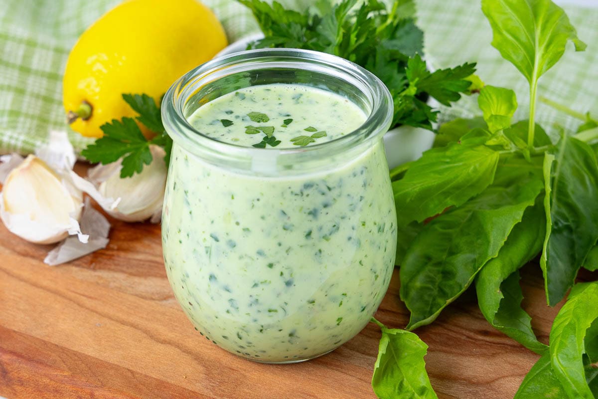 A glass jar filled with green herb sauce sits on a wooden surface, surrounded by fresh basil leaves, parsley, garlic cloves, and a lemon, with a green checkered cloth in the background.