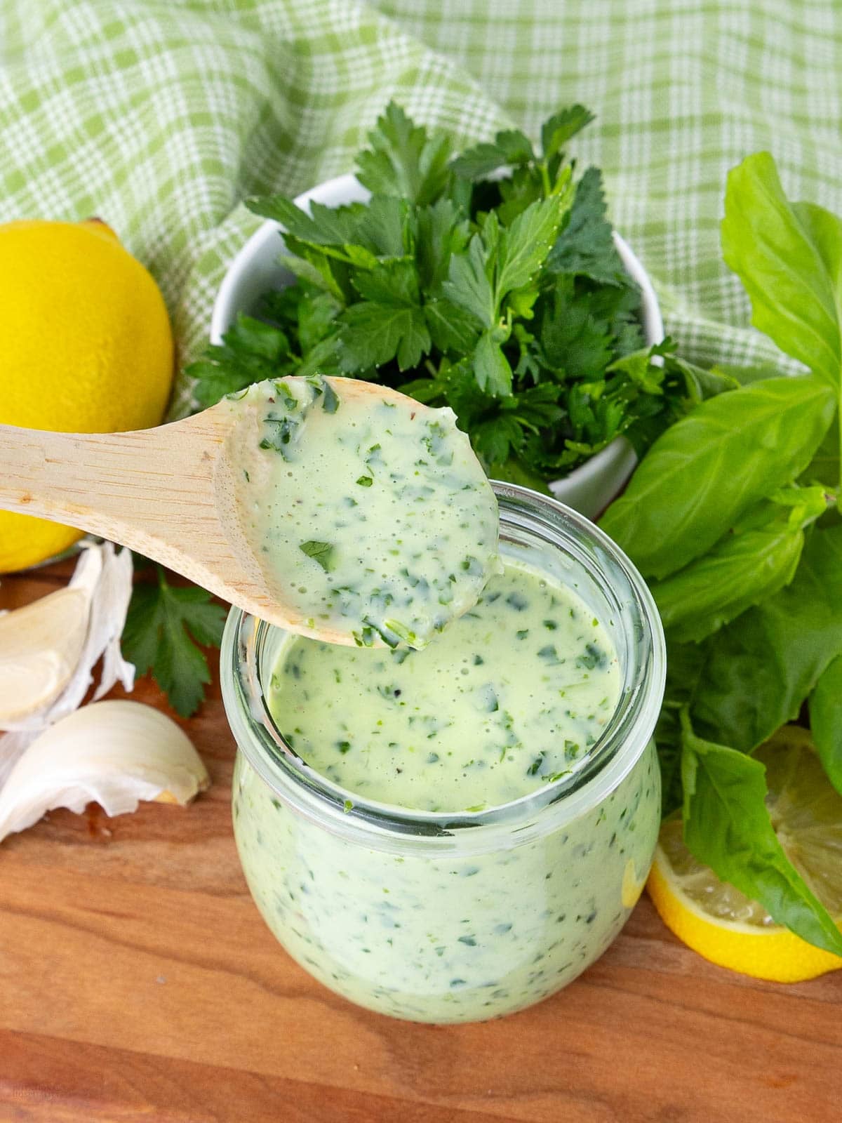 A wooden spoon holds up creamy green herb dressing above a glass jar. Fresh parsley, basil, garlic, whole lemons, and a green checked cloth are arranged on a wooden surface around the jar.