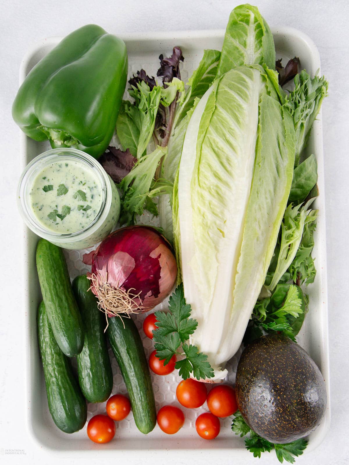 A tray with romaine lettuce, a green bell pepper, mini cucumbers, cherry tomatoes, a red onion, an avocado, mixed salad greens, fresh parsley, and a jar of creamy green salad dressing.