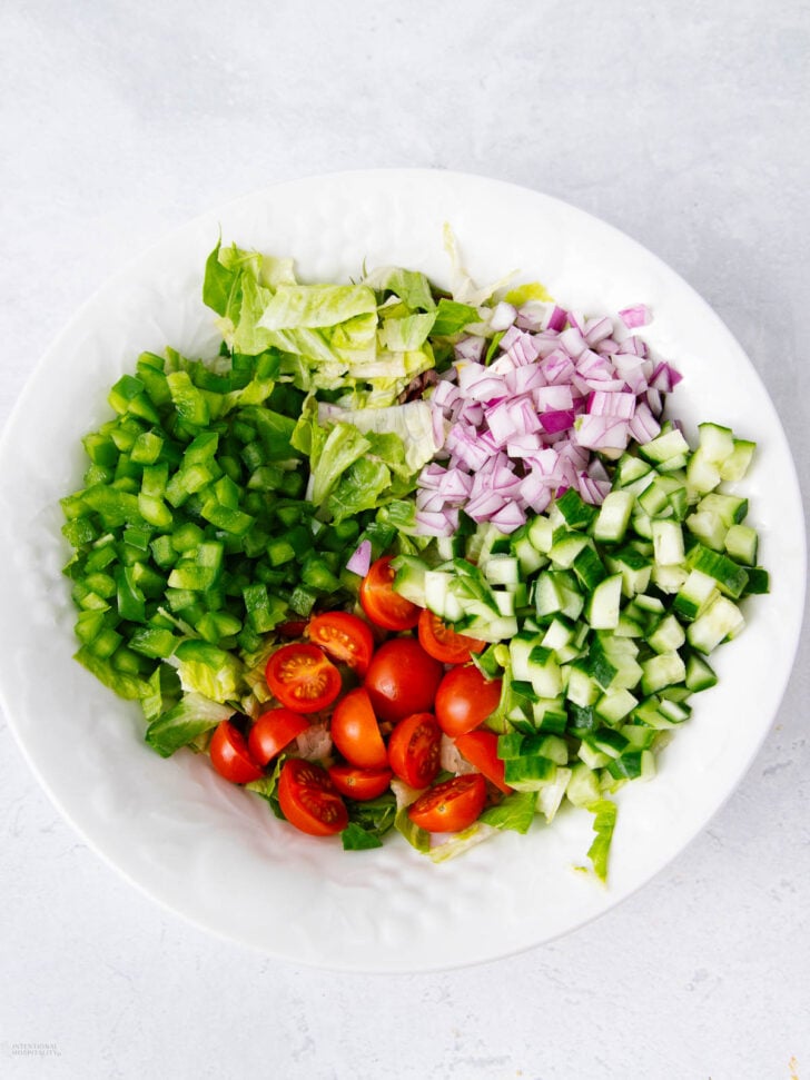 A white bowl contains chopped green bell peppers, red onions, cucumbers, grape tomatoes, and lettuce, arranged in separate sections on a light-colored surface.