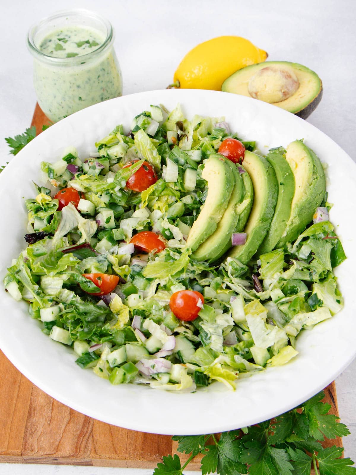 A white bowl filled with chopped salad, cherry tomatoes, cucumber, and topped with sliced avocado. Nearby are a halved avocado, a lemon, and a jar of green dressing on a wooden board.