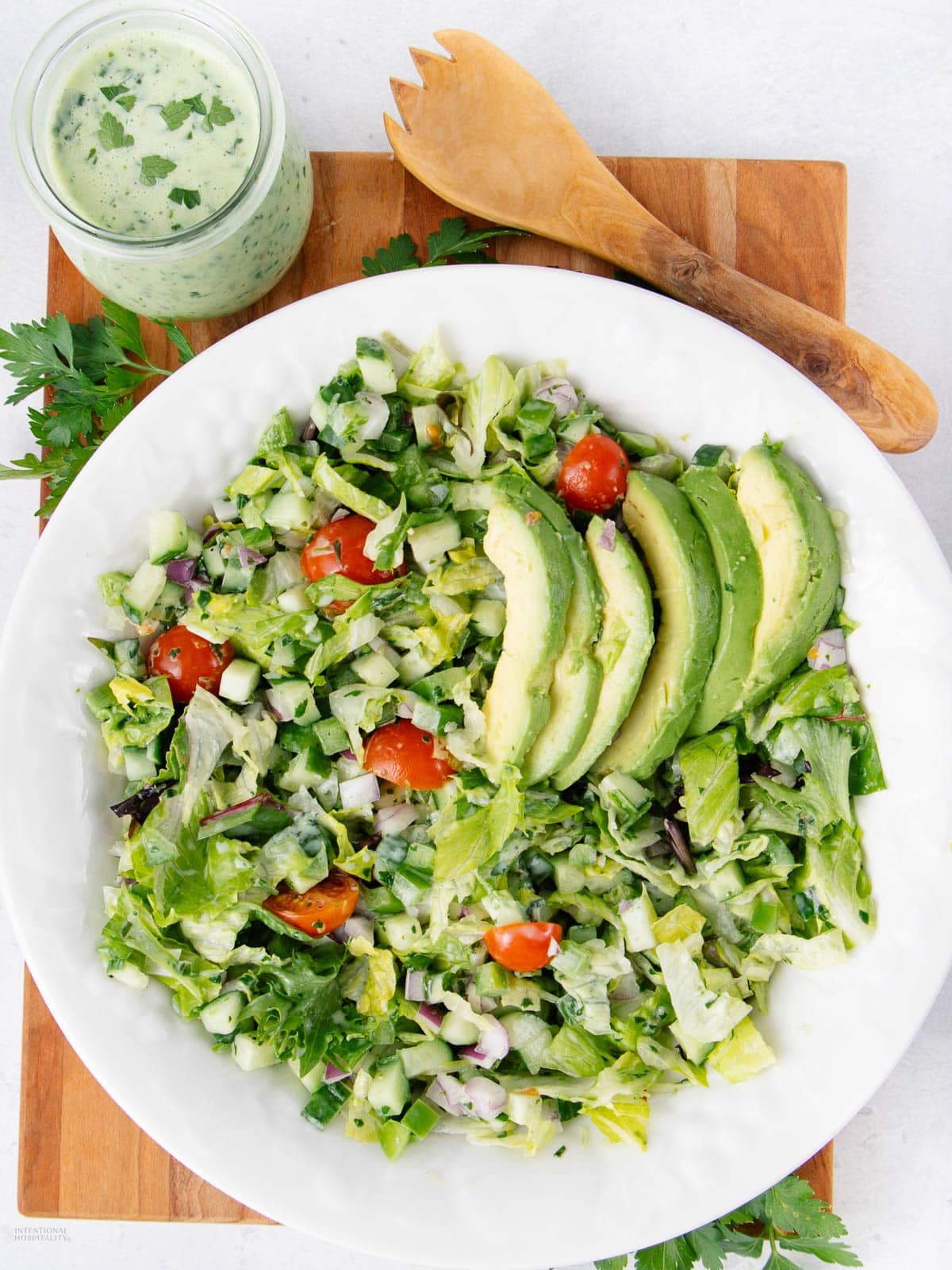 A white bowl of chopped salad with lettuce, tomatoes, cucumber, red onion, and sliced avocado, next to a jar of green dressing and wooden salad utensils on a wooden board.