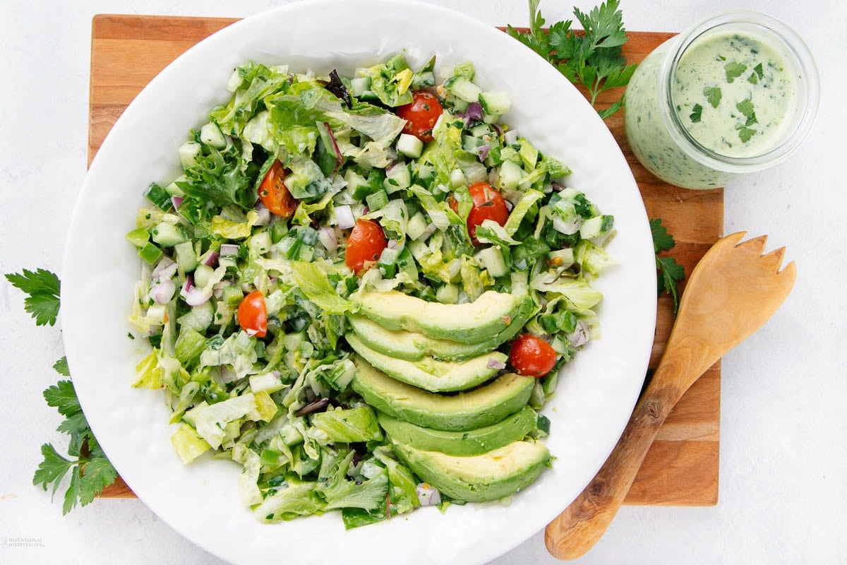 A white bowl with green salad, sliced avocado, cherry tomatoes, cucumbers, and greens, next to a glass jar of creamy herb dressing and a wooden salad utensil on a cutting board.