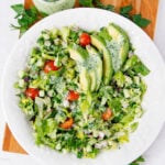 A white plate with a chopped salad of lettuce, cherry tomatoes, cucumber, and red onion, topped with sliced avocado and creamy green herb dressing. Fresh parsley and a jar of dressing are in the background.