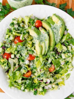 A white plate with a chopped salad of lettuce, cherry tomatoes, cucumber, and red onion, topped with sliced avocado and creamy green herb dressing. Fresh parsley and a jar of dressing are in the background.