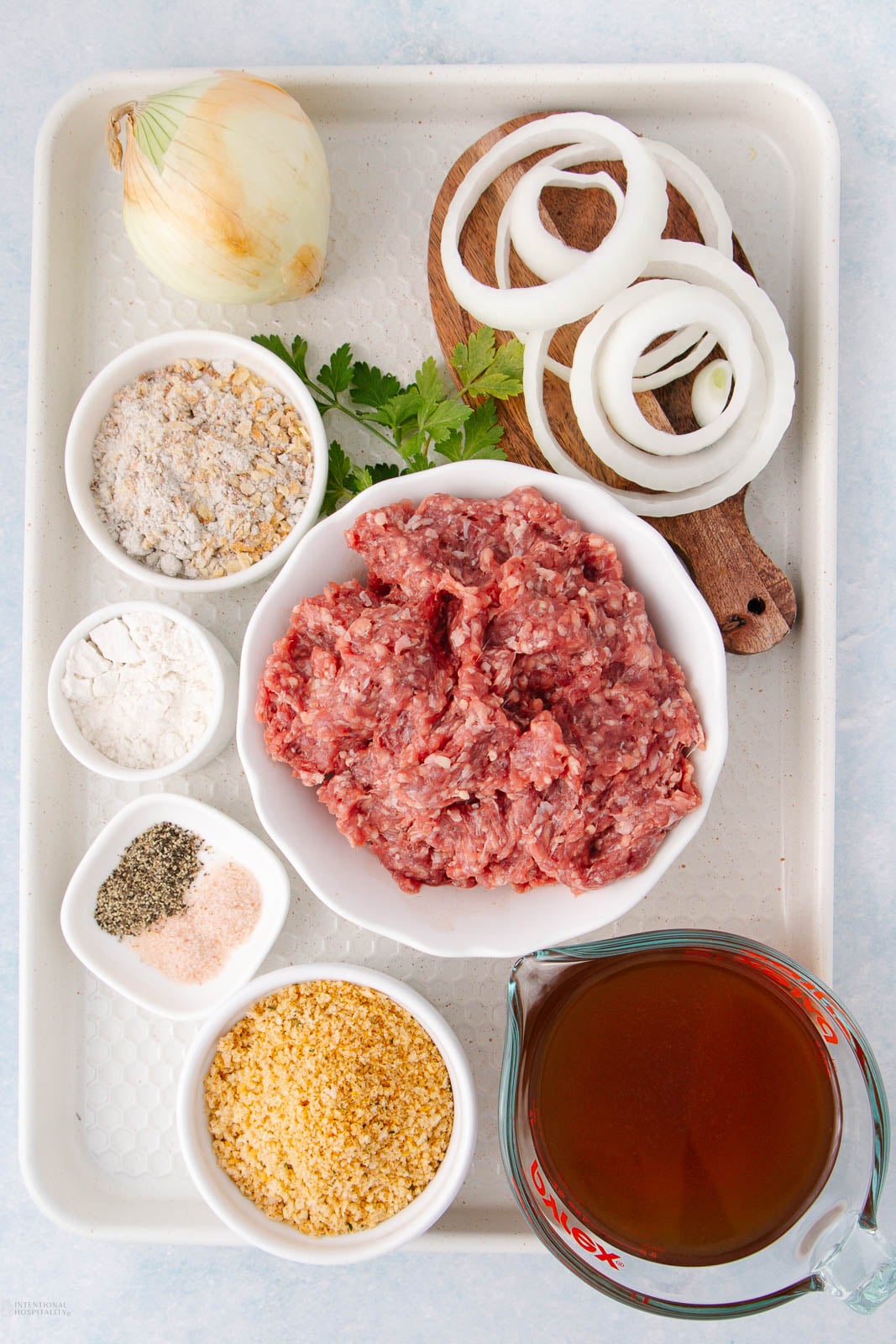 A tray with ground meat in a bowl, onion slices on a small board, a whole onion, parsley, bowls of bread crumbs, flour, seasoning, and a measuring cup of broth.