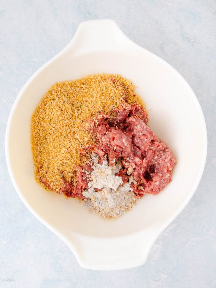 A white mixing bowl containing ground meat, breadcrumbs, and seasoning on a light blue surface, ingredients not yet mixed together.