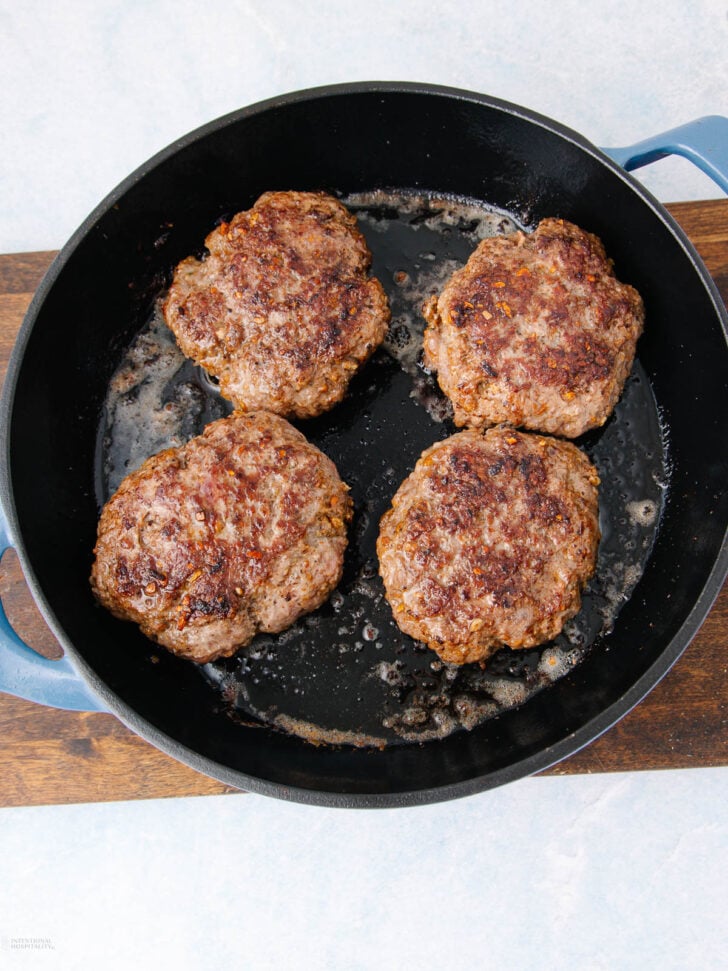 Four browned hamburger patties cooking in a black skillet with some oil, placed on a wooden surface against a light background.