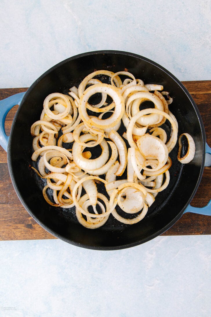 A black skillet with cooked, lightly browned onion rings sits on a wooden board. The skillet has blue handles and is viewed from above against a light blue background.
