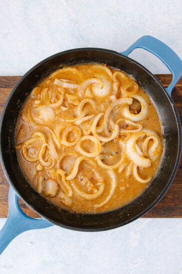 A blue-handled skillet filled with sliced onions simmering in a creamy, brown sauce on a wooden board, viewed from above.