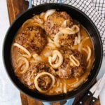 A skillet filled with four hamburger steaks topped with sautéed onions and covered in brown gravy, next to a black and white checkered towel and a knife on a wooden surface.